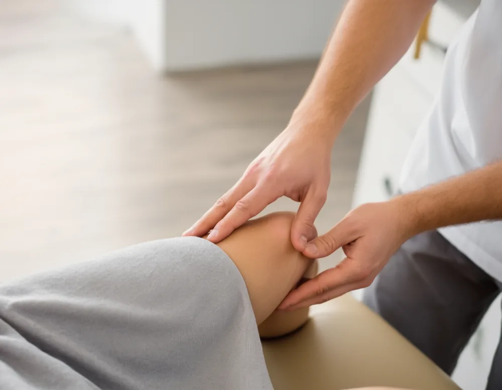 A therapist's hands examining a patient's knee on the treatment table, over a grey linen sheet.