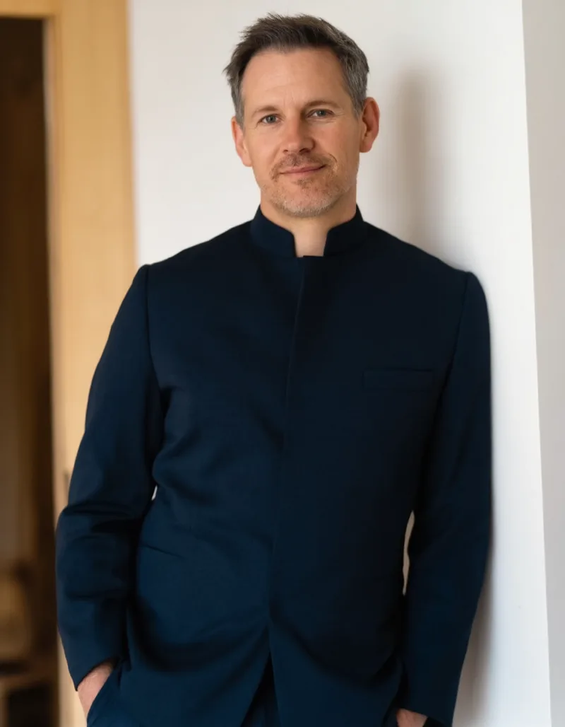 Portrait of Jonas Weisker, physiotherapist, in a charcoal tunic beside a pale oak door frame in the practice.