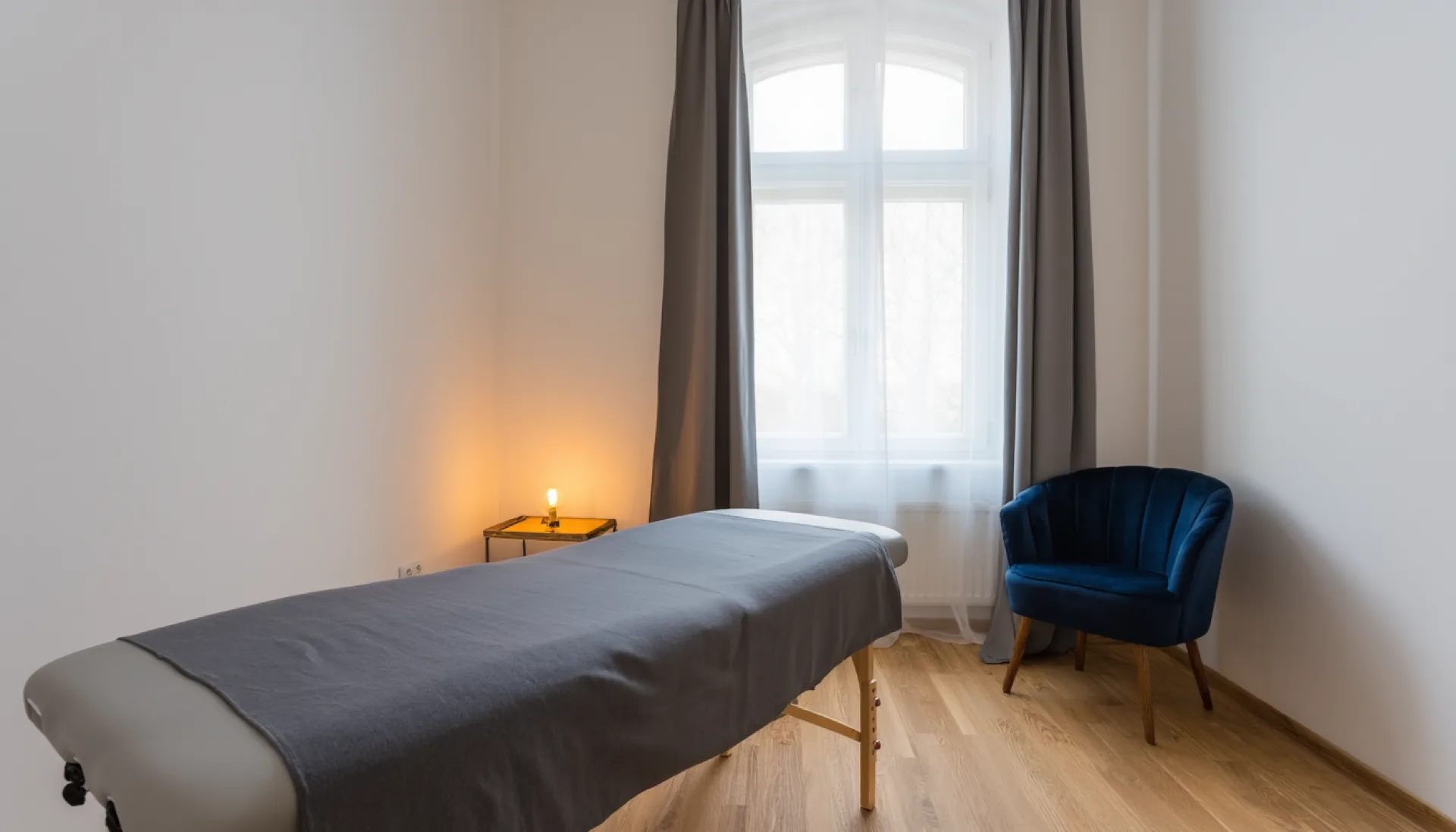 The treatment room at Motus: a treatment table with folded grey linen on a pale oak floor, a linen curtain at the tall window and a low brass lamp.