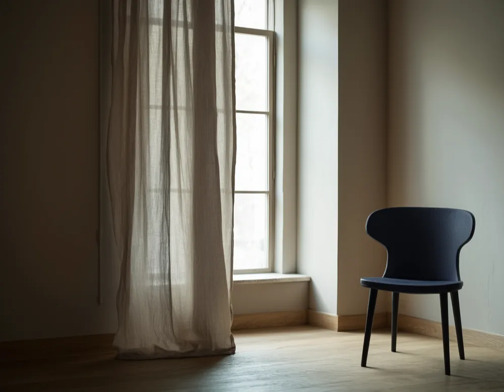 A tall window with a half-drawn unbleached linen curtain, afternoon light on the pale oak floor of the practice.